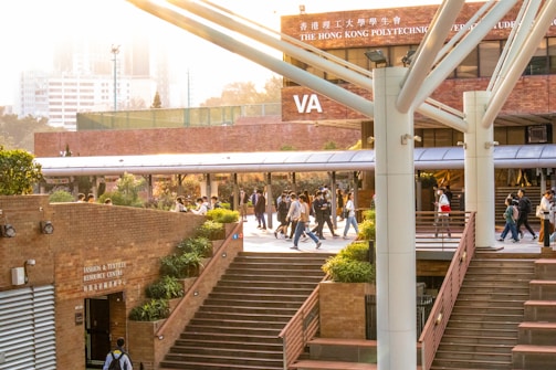 A lively university campus scene with students walking and socializing in a courtyard area. The architecture features modern building elements with brick walls and large support beams. Lush green plants are interspersed around the pathways, contributing to a welcoming atmosphere. The sunlight casts a warm glow over the scene, creating a pleasant and energetic environment.