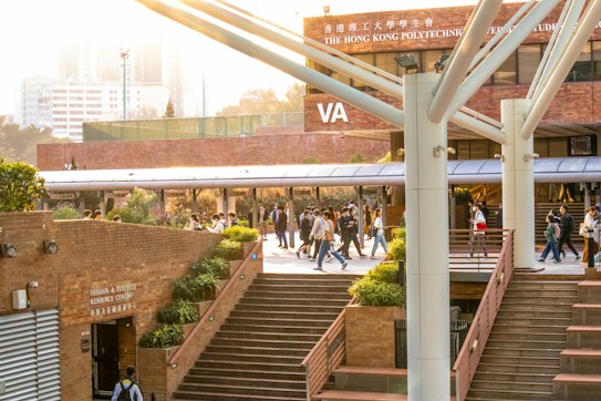 A lively university campus scene with students walking and socializing in a courtyard area. The architecture features modern building elements with brick walls and large support beams. Lush green plants are interspersed around the pathways, contributing to a welcoming atmosphere. The sunlight casts a warm glow over the scene, creating a pleasant and energetic environment.