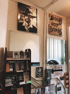 A cozy bookstore interior with a vintage aesthetic. A prominent black-and-white poster of a man wearing a hat hangs on the wall. Next to it, a handmade sign reads 'Books Not Bombs.' Below, a wooden shelf displays various books. Another poster encourages literacy with 'Learn to Read.' The walls have light panels and other small posters are present.