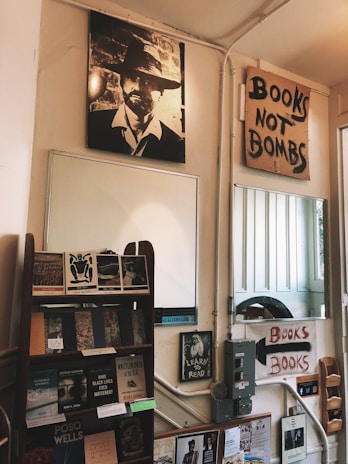 A cozy bookstore interior with a vintage aesthetic. A prominent black-and-white poster of a man wearing a hat hangs on the wall. Next to it, a handmade sign reads 'Books Not Bombs.' Below, a wooden shelf displays various books. Another poster encourages literacy with 'Learn to Read.' The walls have light panels and other small posters are present.