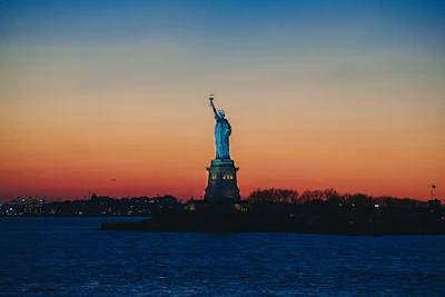 The Statue of Liberty with Ellis Island in the background at sunset.