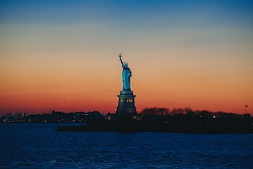 The Statue of Liberty stands prominently against a vibrant sunset backdrop, with deep orange and blue hues blending seamlessly in the sky. The silhouette of the cityscape is faintly visible across the water.