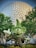 A large, green topiary sculpture of a cartoon character stands in front of a futuristic geodesic dome structure. The topiary is surrounded by colorful flowers and lush green trees, creating a vibrant garden scene.