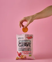 A hand holds a single round chip above a pink bag of The Organic Crave protein chips. The flavor of the chips is paprika and onion, as indicated on the package. The scene has a soft pink background that matches the color of the packaging.