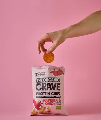A hand holds a single round chip above a pink bag of The Organic Crave protein chips. The flavor of the chips is paprika and onion, as indicated on the package. The scene has a soft pink background that matches the color of the packaging.