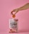 A hand holds a single round chip above a pink bag of The Organic Crave protein chips. The flavor of the chips is paprika and onion, as indicated on the package. The scene has a soft pink background that matches the color of the packaging.