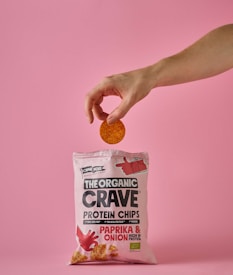 A hand holds a single round chip above a pink bag of The Organic Crave protein chips. The flavor of the chips is paprika and onion, as indicated on the package. The scene has a soft pink background that matches the color of the packaging.