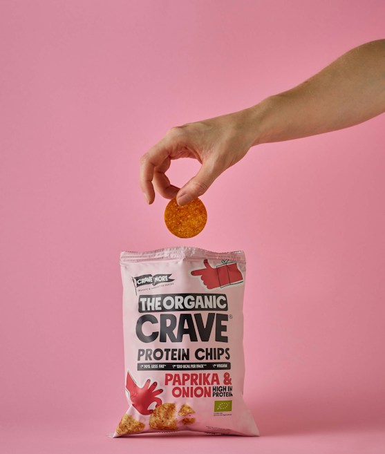 A hand holds a single round chip above a pink bag of The Organic Crave protein chips. The flavor of the chips is paprika and onion, as indicated on the package. The scene has a soft pink background that matches the color of the packaging.