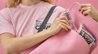 A person wearing a pink T-shirt is holding a pink bag while placing snack bars inside. The snack bars have colorful packaging and the text 'The Organic Crave' visible on them.