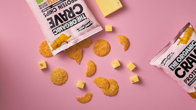 A flat lay of snack items against a pink background, featuring two open bags of protein chips with pieces scattered around. Several small cubes and a larger slice of cheese accompany the chips.