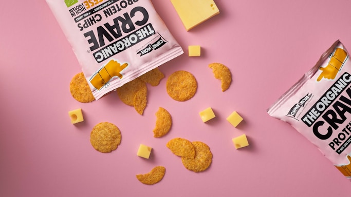 A flat lay of snack items against a pink background, featuring two open bags of protein chips with pieces scattered around. Several small cubes and a larger slice of cheese accompany the chips.