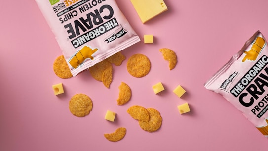 A flat lay of snack items against a pink background, featuring two open bags of protein chips with pieces scattered around. Several small cubes and a larger slice of cheese accompany the chips.