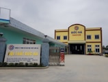 A large building with the words 'Đức Hà Furniture' displayed prominently at the top. The structure has a yellow facade with brown trim and blue window frames. In front, a signboard displays additional information about the business in Vietnamese, with contact details and a website. The entrance is wide and gated, flanked by decorative stone pillars with spherical lamps on top. The area is open and well-maintained.