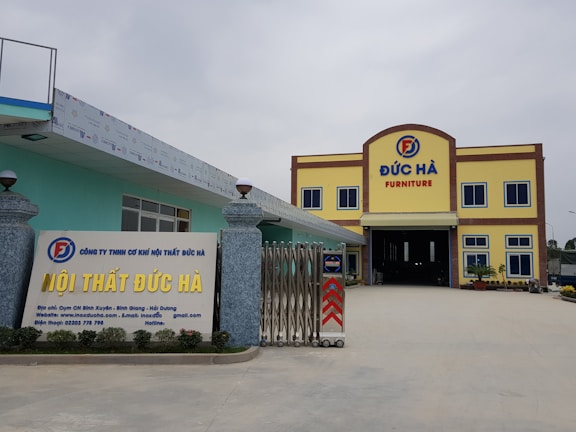 A large building with the words 'Đức Hà Furniture' displayed prominently at the top. The structure has a yellow facade with brown trim and blue window frames. In front, a signboard displays additional information about the business in Vietnamese, with contact details and a website. The entrance is wide and gated, flanked by decorative stone pillars with spherical lamps on top. The area is open and well-maintained.