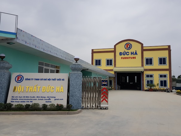 A large building with the words 'Đức Hà Furniture' displayed prominently at the top. The structure has a yellow facade with brown trim and blue window frames. In front, a signboard displays additional information about the business in Vietnamese, with contact details and a website. The entrance is wide and gated, flanked by decorative stone pillars with spherical lamps on top. The area is open and well-maintained.