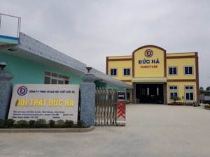 A large building with the words 'Đức H&agrave; Furniture' displayed prominently at the top. The structure has a yellow facade with brown trim and blue window frames. In front, a signboard displays additional information about the business in Vietnamese, with contact details and a website. The entrance is wide and gated, flanked by decorative stone pillars with spherical lamps on top. The area is open and well-maintained.
