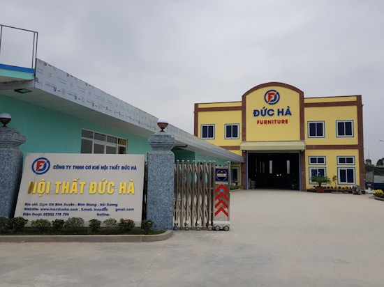 A large building with the words 'Đức H&agrave; Furniture' displayed prominently at the top. The structure has a yellow facade with brown trim and blue window frames. In front, a signboard displays additional information about the business in Vietnamese, with contact details and a website. The entrance is wide and gated, flanked by decorative stone pillars with spherical lamps on top. The area is open and well-maintained.