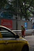 A person wearing an orange uniform and a reflective vest stands on a street, holding a cleaning tool. A yellow taxi passes by in the foreground. The setting includes a metal fence with signage in Chinese and green trees in the background.