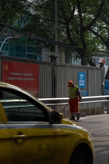 A person wearing an orange uniform and a reflective vest stands on a street, holding a cleaning tool. A yellow taxi passes by in the foreground. The setting includes a metal fence with signage in Chinese and green trees in the background.