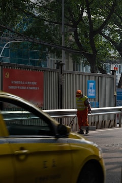 A person wearing an orange uniform and a reflective vest stands on a street, holding a cleaning tool. A yellow taxi passes by in the foreground. The setting includes a metal fence with signage in Chinese and green trees in the background.