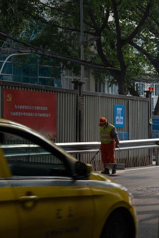 A person wearing an orange uniform and a reflective vest stands on a street, holding a cleaning tool. A yellow taxi passes by in the foreground. The setting includes a metal fence with signage in Chinese and green trees in the background.