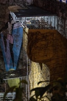 A large, illuminated climbing wall with vividly colored grips is set against a rugged, natural rock surface. Overhead, there is a metal structure with a small, roofed platform where a person is visible. The scene includes elements of nature, such as ivy or plant life, climbing the surface of both the wall and the rock face.