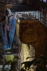 A large, illuminated climbing wall with vividly colored grips is set against a rugged, natural rock surface. Overhead, there is a metal structure with a small, roofed platform where a person is visible. The scene includes elements of nature, such as ivy or plant life, climbing the surface of both the wall and the rock face.
