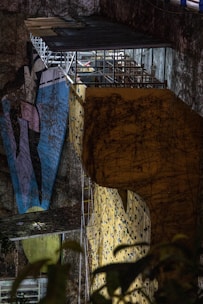 A large, illuminated climbing wall with vividly colored grips is set against a rugged, natural rock surface. Overhead, there is a metal structure with a small, roofed platform where a person is visible. The scene includes elements of nature, such as ivy or plant life, climbing the surface of both the wall and the rock face.