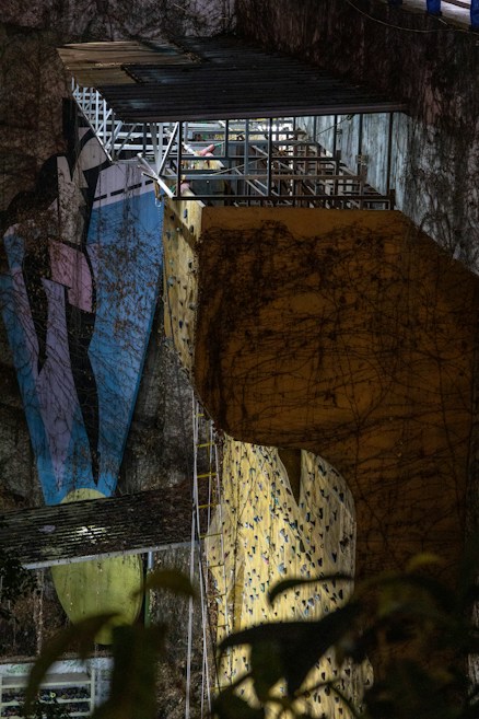 A large, illuminated climbing wall with vividly colored grips is set against a rugged, natural rock surface. Overhead, there is a metal structure with a small, roofed platform where a person is visible. The scene includes elements of nature, such as ivy or plant life, climbing the surface of both the wall and the rock face.