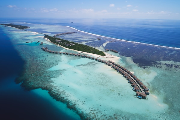 An aerial view of a tropical island surrounded by clear turquoise waters and coral reefs. Overwater bungalows extend into the sea, connected by a series of walkways. Lush green vegetation covers parts of the island, and there are sandy beaches. The sky is clear with a few small clouds, and the ocean stretches out to the horizon.