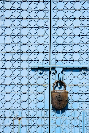 A close-up view of a blue metal gate with an intricate geometric pattern. The gate features a large, old-fashioned padlock that is prominently placed in the center, adding a sense of security and closure.