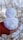 A handmade snowman figure is cradled in a person's hand, adorned with simple twig arms and facial features. The background is softly blurred with dry, brown branches. The snowman has a slightly asymmetrical shape, giving it a charmingly rustic appearance.