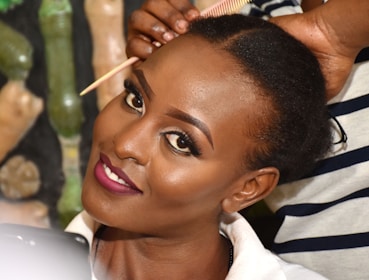 A person with well-defined makeup and vibrant lipstick is smiling while another individual styles their hair using a comb. The background consists of various blurred objects in neutral and green shades.