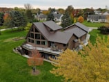 A large, modern house with a unique architectural design featuring multiple gables and a spacious balcony. The house is surrounded by well-manicured lawns and numerous trees, displaying autumn foliage. The neighborhood consists of similarly styled homes, set against a backdrop of mixed forest and open skies.