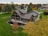 A large, modern house with a unique architectural design featuring multiple gables and a spacious balcony. The house is surrounded by well-manicured lawns and numerous trees, displaying autumn foliage. The neighborhood consists of similarly styled homes, set against a backdrop of mixed forest and open skies.