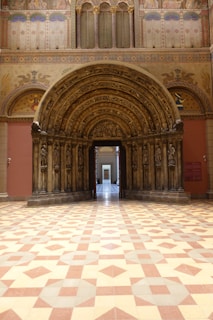 A grand, ornately carved stone archway that serves as an entrance in a richly decorated hall. The archway is adorned with sculptures of figures and detailed patterns. The walls display intricate paintings and designs featuring rich colors and gold elements. The patterned floor complements the elaborate architecture, and a doorway provides a view into another space.