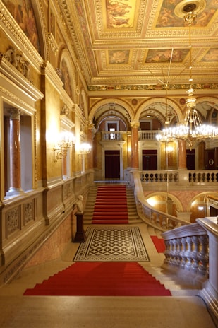 A grand, opulent interior featuring a decorative staircase adorned with red carpets. The walls and ceiling are intricately designed with gold accents, detailed paintings, and ornate chandeliers. There are multiple archways, columns, and luxurious light fixtures illuminating the space.
