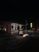 A dimly lit gas station at night featuring a prominent sign displaying fuel options. The area is covered with snow, reflecting some of the light from the station. A few cars are parked under the canopies, and the street beyond is faintly visible.