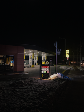 A dimly lit gas station at night featuring a prominent sign displaying fuel options. The area is covered with snow, reflecting some of the light from the station. A few cars are parked under the canopies, and the street beyond is faintly visible.