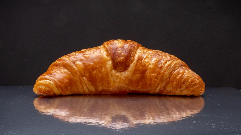 Close-up of a freshly baked golden croissant resting on a beige linen cloth with a dark pink background.
