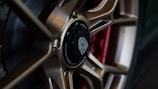 Close-up view of a stylish car wheel with a metallic alloy rim and a central black hub featuring detailed engravings. The wheel design is intricate, showcasing a blend of bronze tones against a red component in the background.