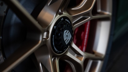 Close-up view of a stylish car wheel with a metallic alloy rim and a central black hub featuring detailed engravings. The wheel design is intricate, showcasing a blend of bronze tones against a red component in the background.