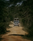A rugged off-road vehicle is traveling along a dirt path surrounded by dense foliage and trees. The path is uneven, emphasizing a sense of adventure. The vehicle has a roof rack and appears to be a safari or tour jeep with passengers visible inside.