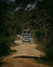 A rugged off-road vehicle is traveling along a dirt path surrounded by dense foliage and trees. The path is uneven, emphasizing a sense of adventure. The vehicle has a roof rack and appears to be a safari or tour jeep with passengers visible inside.