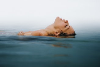 A person is floating peacefully on their back in calm water, with eyes closed and head partially submerged. The horizon blends seamlessly into the sky, creating a tranquil and serene atmosphere.