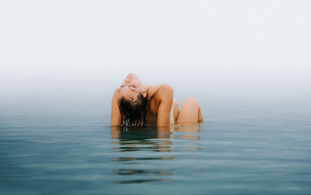 A person is submerged in calm water, leaning back with eyes closed and arms resting on the head. The background is a soft blend of sky and water, creating a serene and peaceful atmosphere.