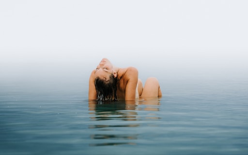 A person is submerged in calm water, leaning back with eyes closed and arms resting on the head. The background is a soft blend of sky and water, creating a serene and peaceful atmosphere.