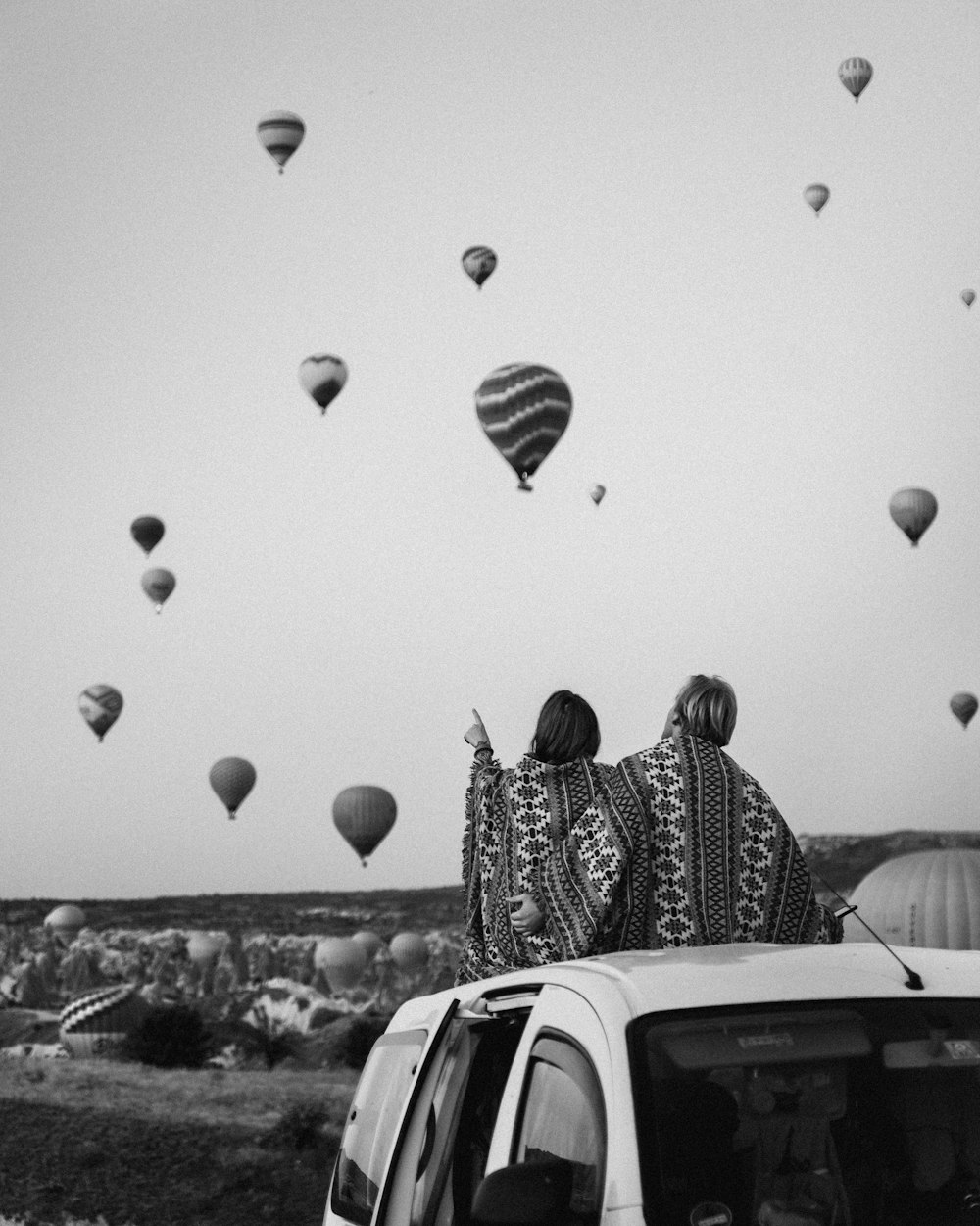 two people sitting on the roof of a car watching hot air balloons in the sky
