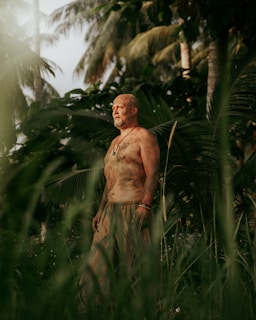 A bare-chested man covered in dirt stands in a jungle-like environment with lush green palm trees and foliage surrounding him. He appears to be deep in thought or contemplation.