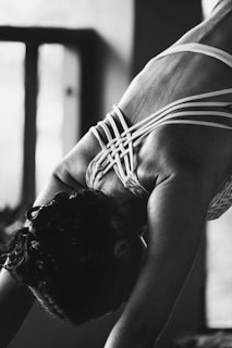 A person is engaged in a yoga or stretching pose, focusing on their back muscles. The intricate design of the straps on their clothing contrasts with the smooth skin. The image is in black and white, providing a dramatic and intimate atmosphere.